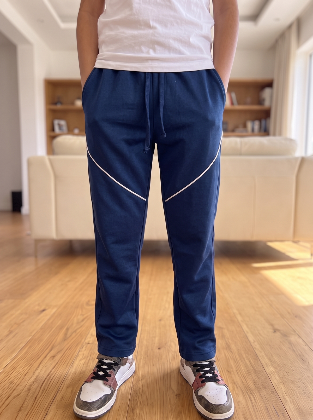 A person standing indoors wearing navy blue sweatpants with white diagonal stripe details on the thighs, a white t-shirt, and white sneakers, photographed from the waist down in a living room setting.