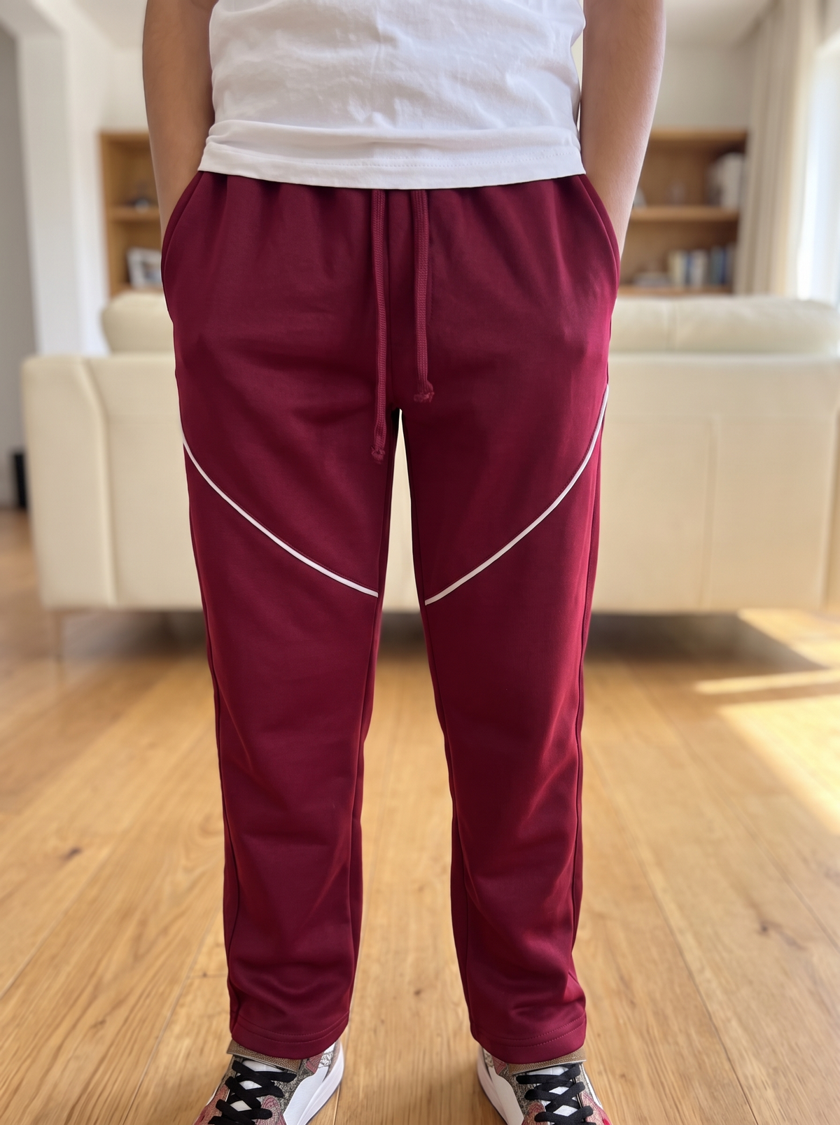 A person wearing burgundy jogger-style pants with white diagonal stripe accents on the thighs, paired with a white t-shirt and black-and-white sneakers, standing in a living room with wooden floors and a beige sofa in the background.