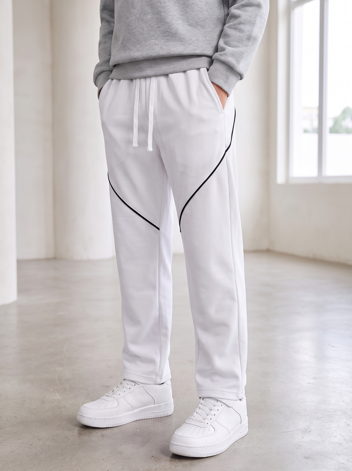 White sweatpants with black diagonal piping details, worn by a model in a gray sweatshirt and white sneakers in a bright, modern room.