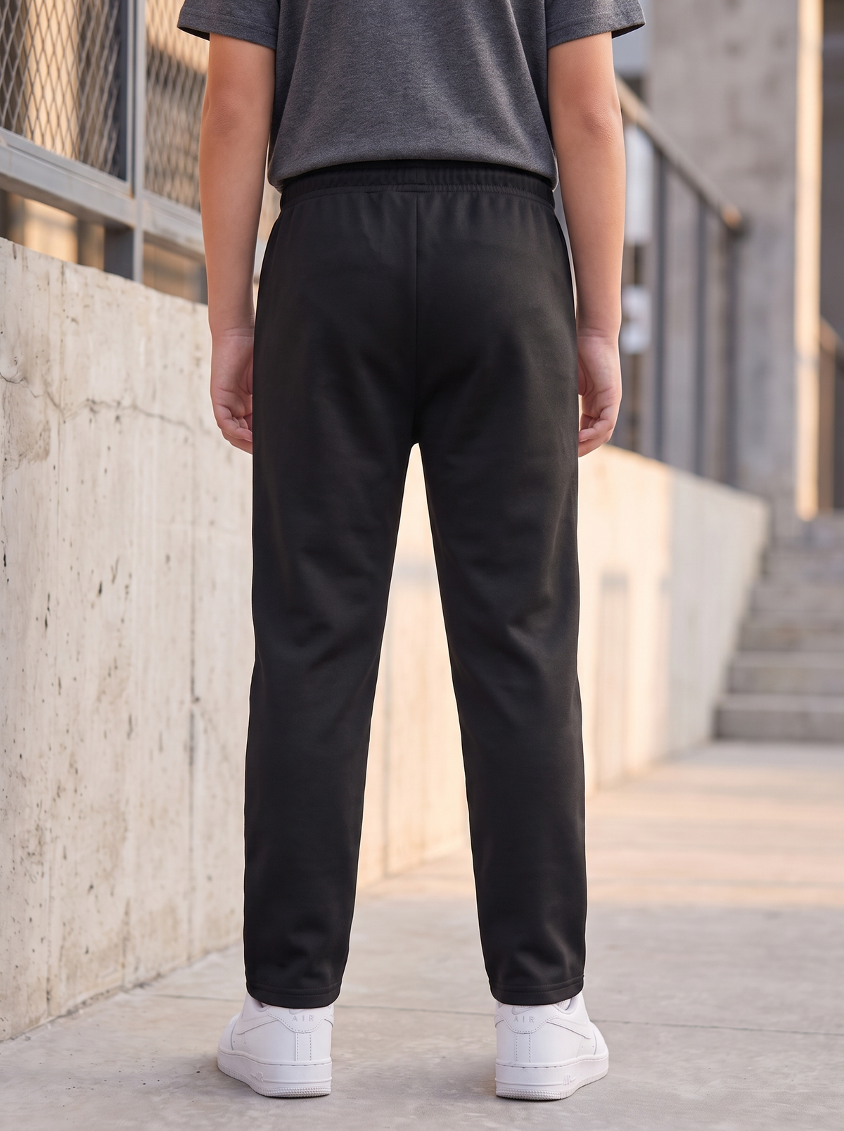 A person viewed from behind wearing black jogger-style pants, a dark gray t-shirt, and white sneakers, standing outdoors against a concrete wall and staircase.