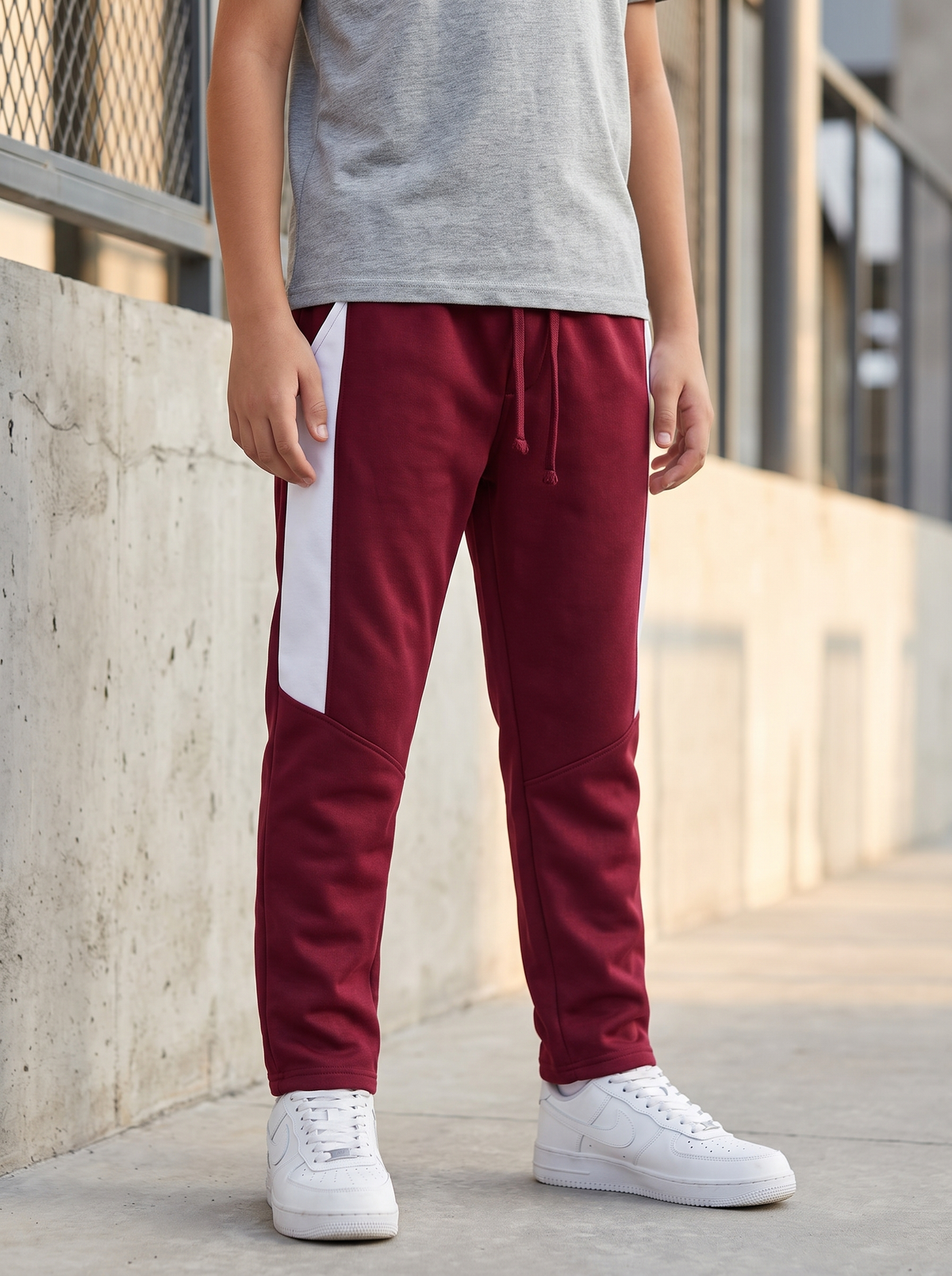 A child wearing burgundy sweatpants with a white side stripe, a gray t-shirt, and white sneakers, standing outdoors next to a concrete wall.