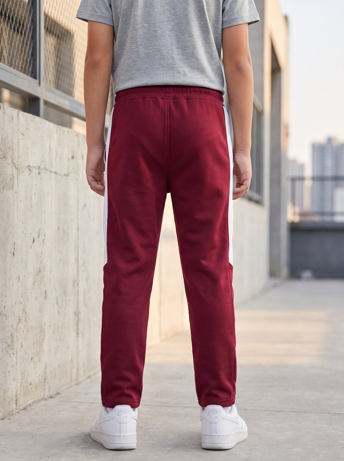 A person viewed from behind wearing burgundy track pants with white side stripes, a gray t-shirt, and white sneakers, standing outdoors on a concrete walkway.