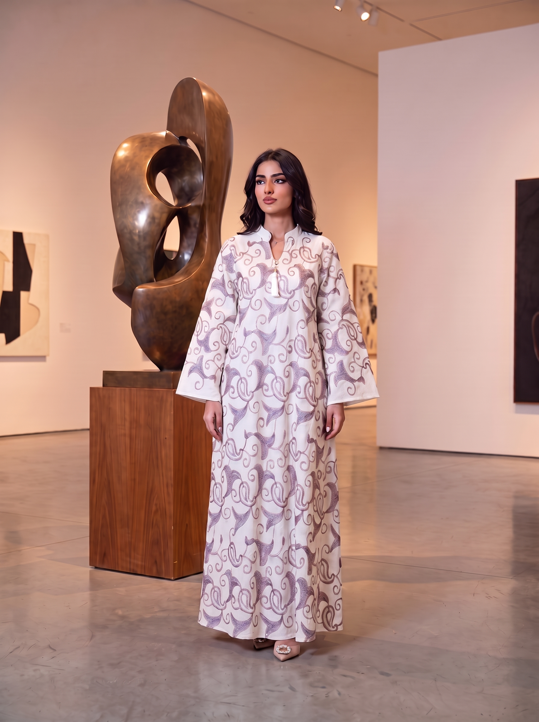 A woman stands in a gallery wearing a long white abaya with a lavender-purple abstract swirl pattern. She is positioned in front of a large bronze abstract sculpture on a wooden pedestal.