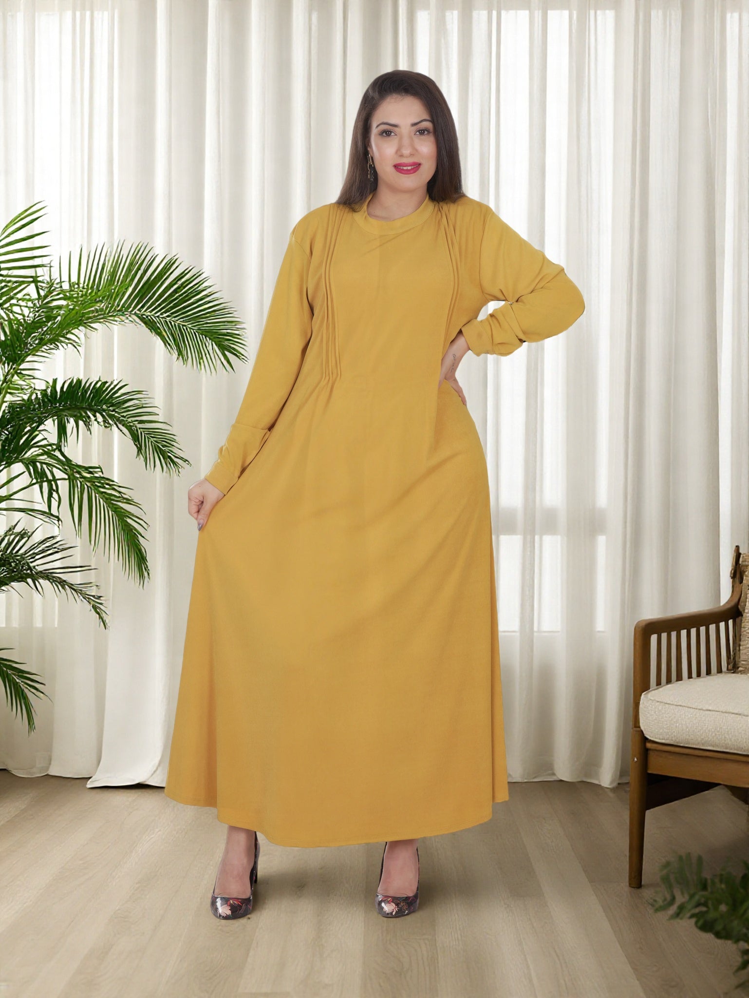 Woman wearing a mustard yellow dress standing in a room with white curtains and a plant.