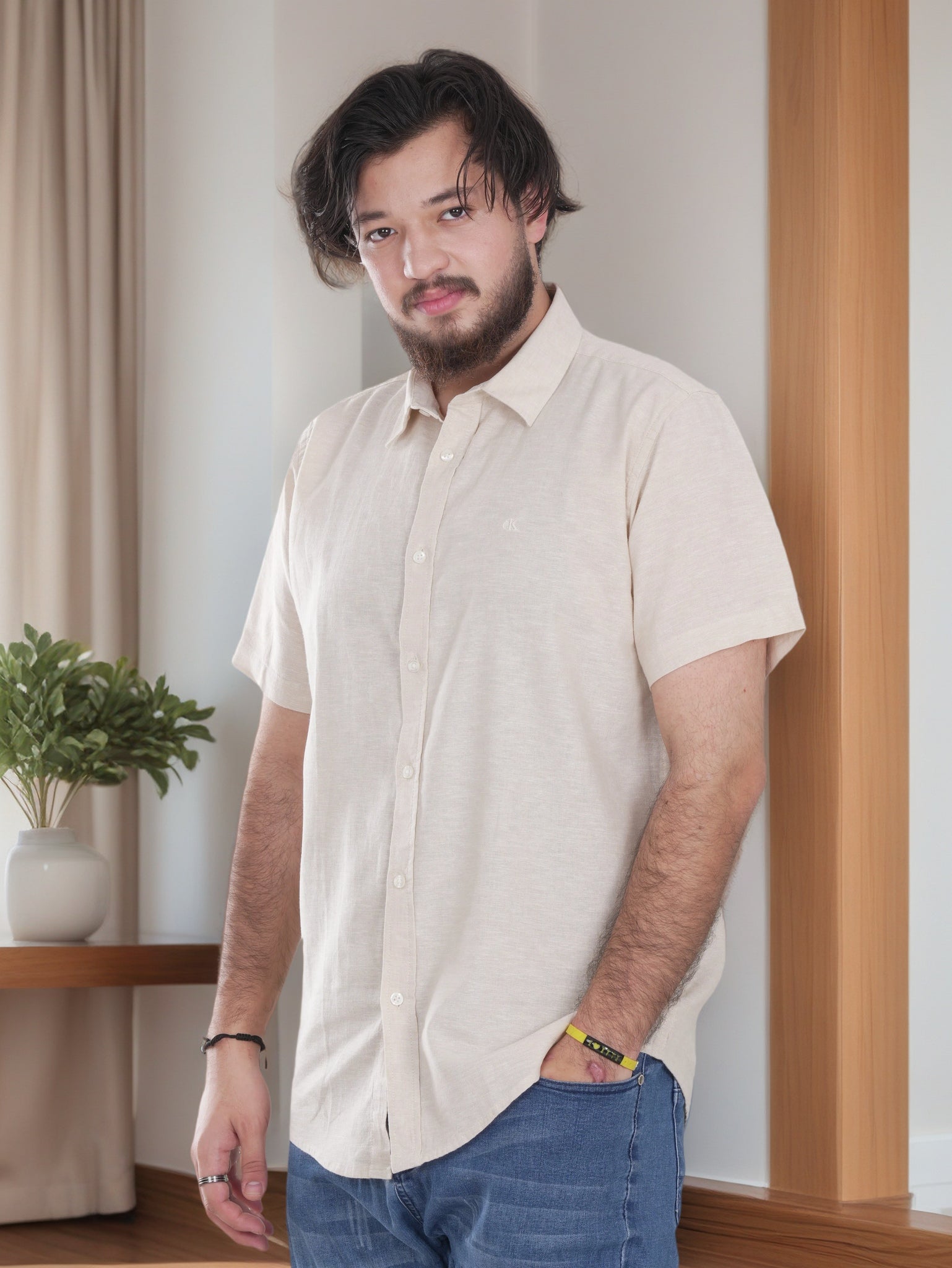 Man wearing a beige shirt and blue jeans standing indoors with a plant and wooden shelf in the background.