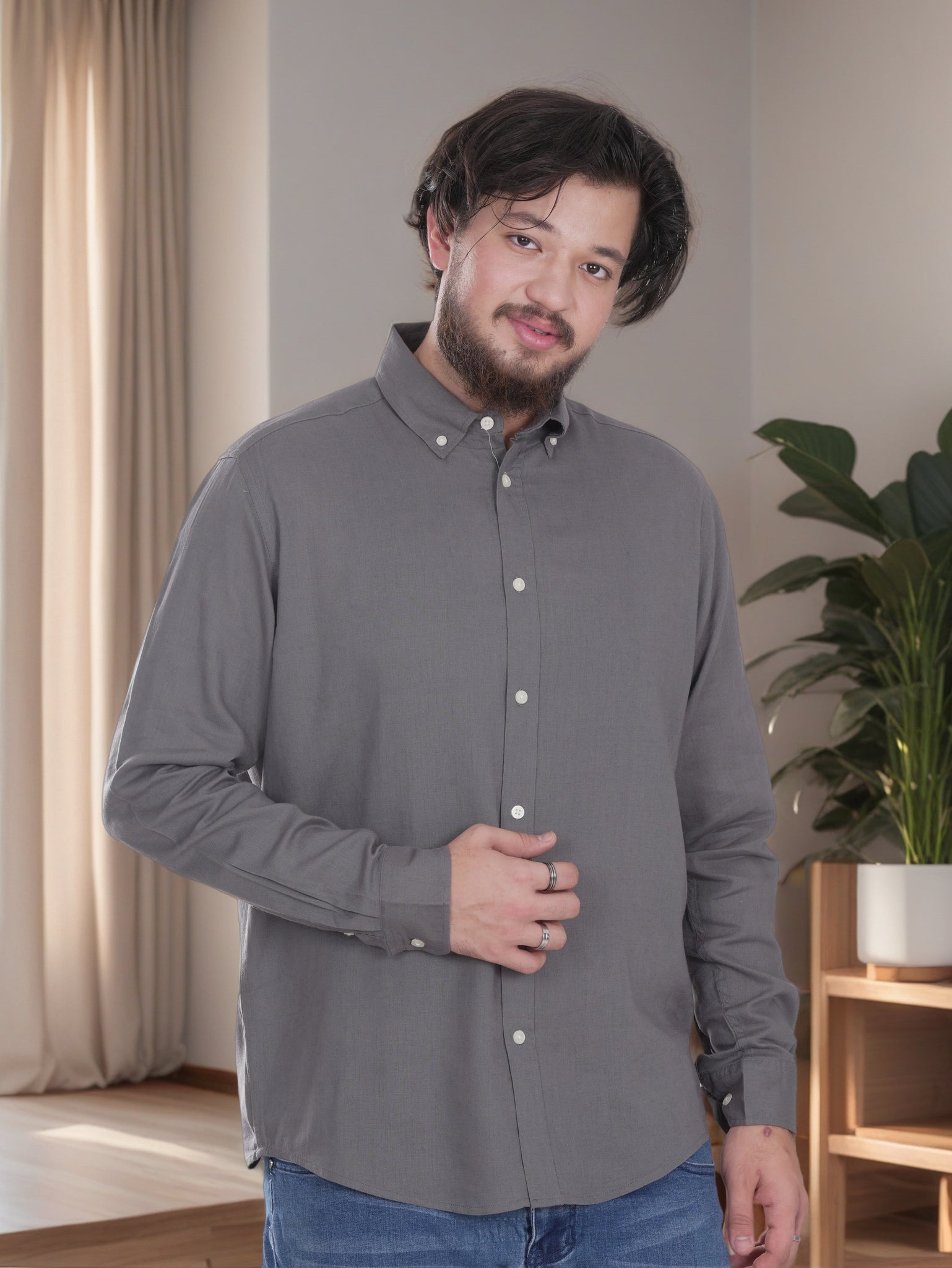 Man wearing a gray shirt standing in a room with a plant and curtains.