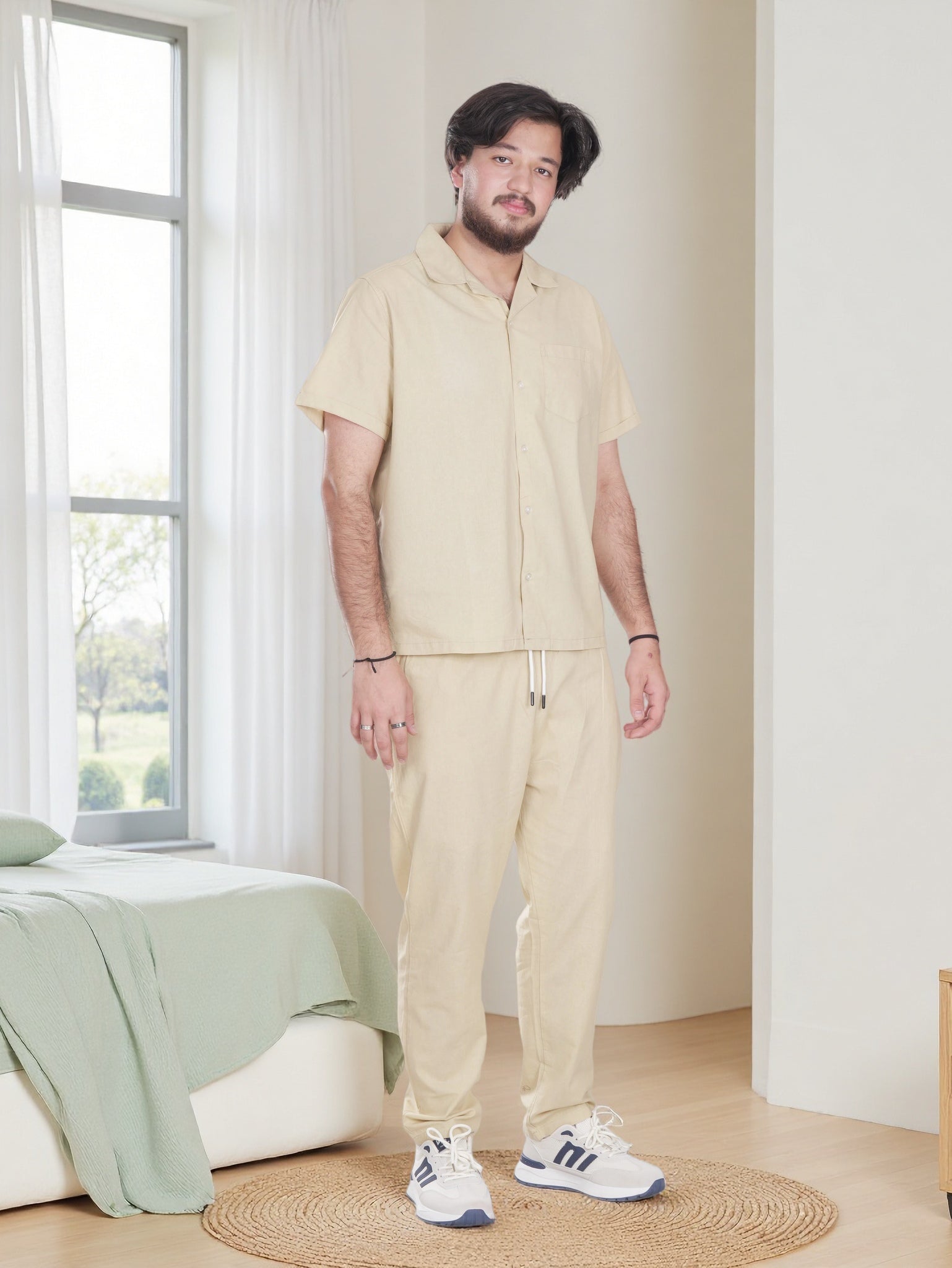 Man wearing a beige outfit standing in a room with a bed and window.