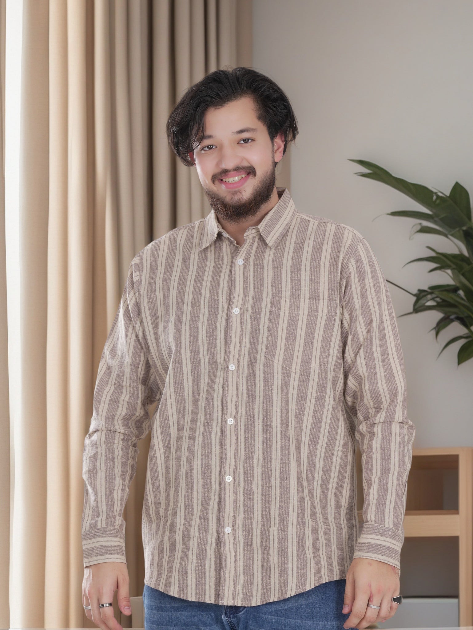 Man wearing a striped shirt standing indoors with curtains and a plant in the background