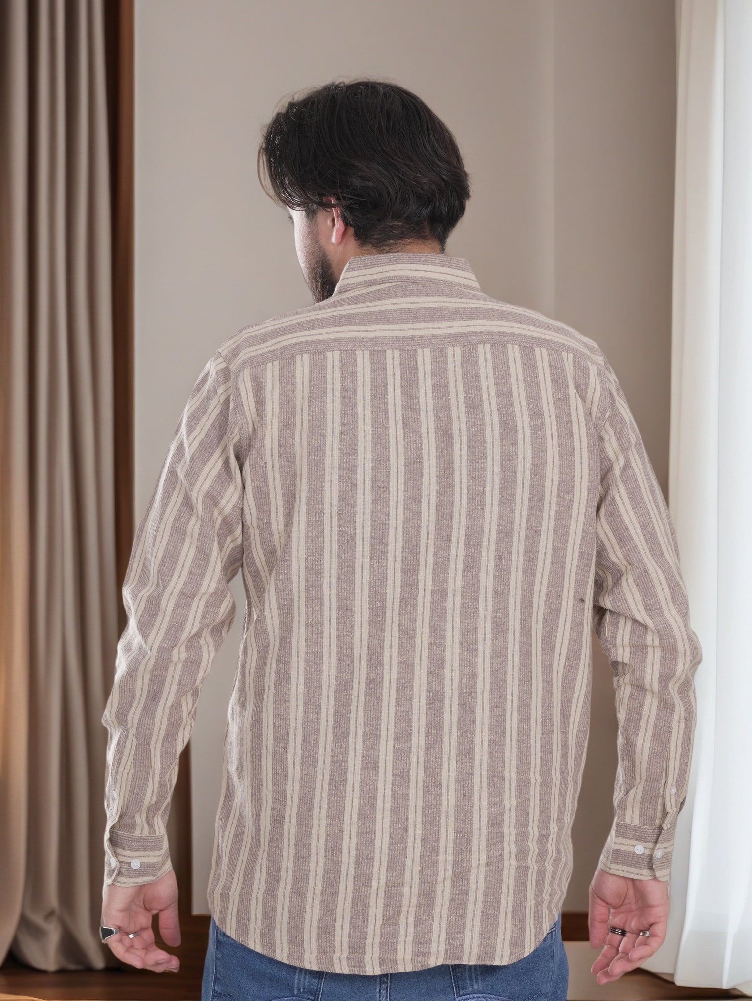 Man wearing a striped shirt standing in a room with curtains.