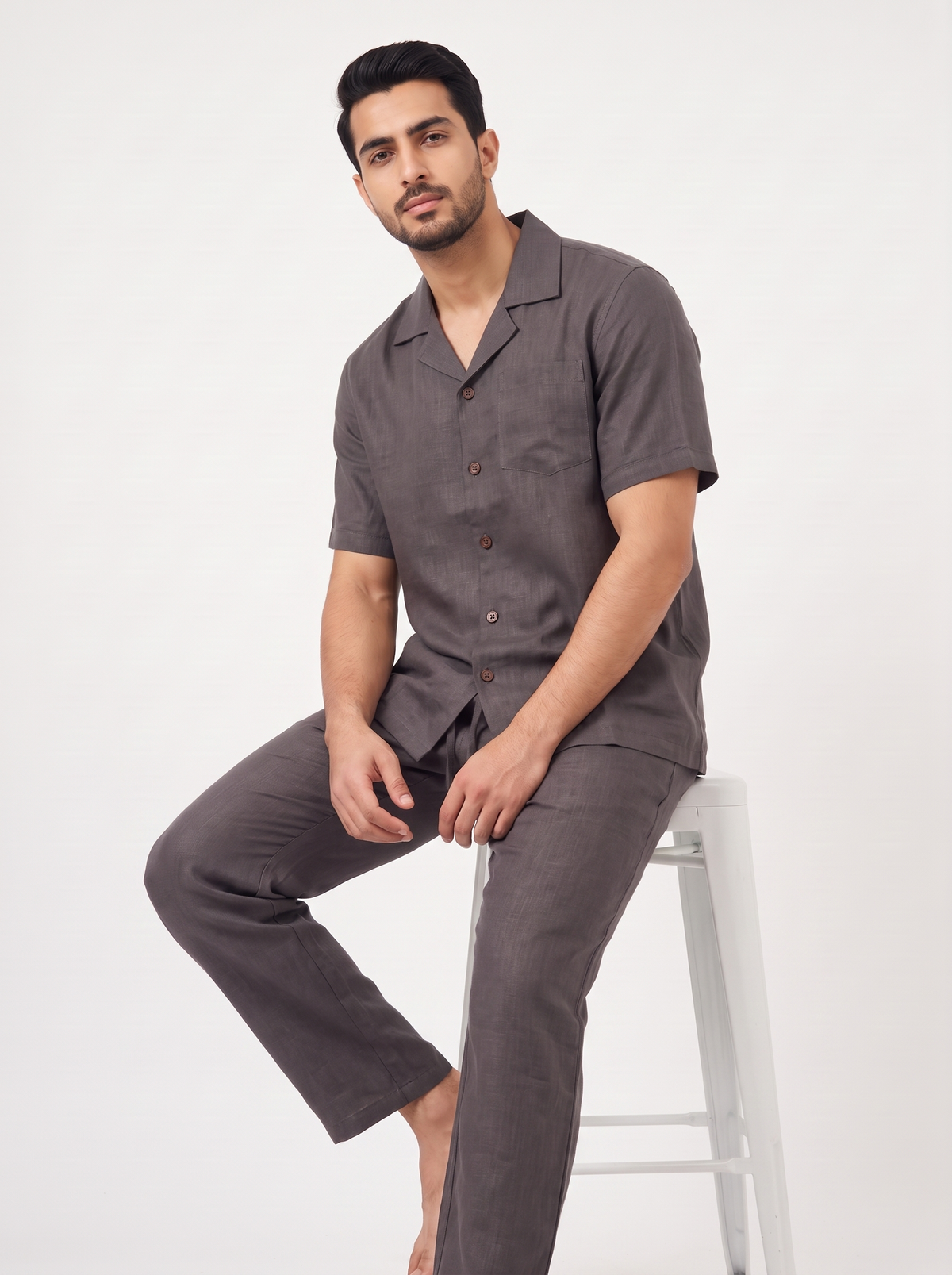 A man wearing a matching dark gray short-sleeve button-up shirt and trousers pajama set, sitting on a white stool against a plain white background.