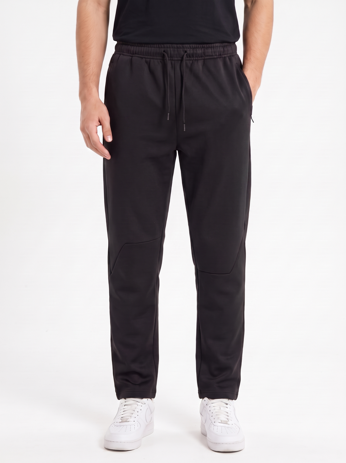 A person wearing black jogger-style pants with a drawstring waist, shown from the chest down against a clean white studio background. The pants have side pockets and a straight-leg fit, paired with white sneakers.