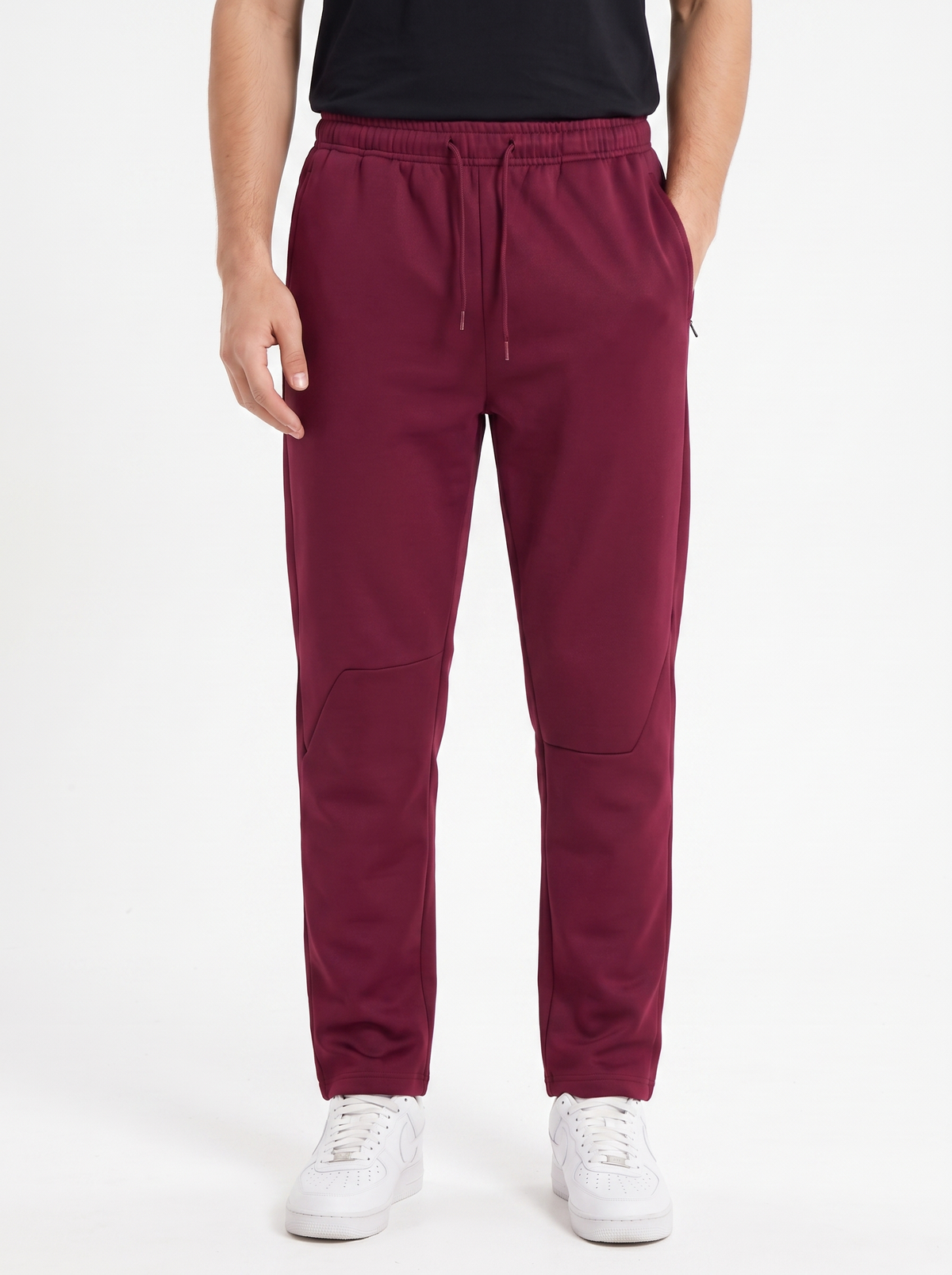 A person wearing burgundy jogger sweatpants with a drawstring waist, paired with a black top and white sneakers, shown from the waist down on a light gray studio background.