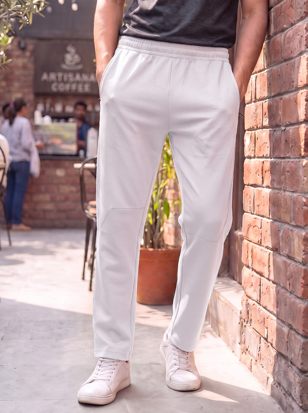 A person standing outdoors in front of a brick wall wearing light gray drawstring sweatpants, a dark gray t-shirt, and white sneakers. The background shows a cafe counter with a sign reading Artisanal Coffee.