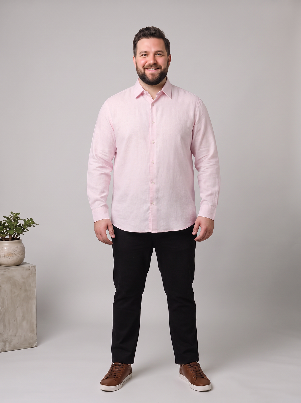 A man wearing a light pink long-sleeve button-down shirt, black trousers, and brown sneakers, standing against a light gray studio background with a small potted plant on a concrete block to the left.