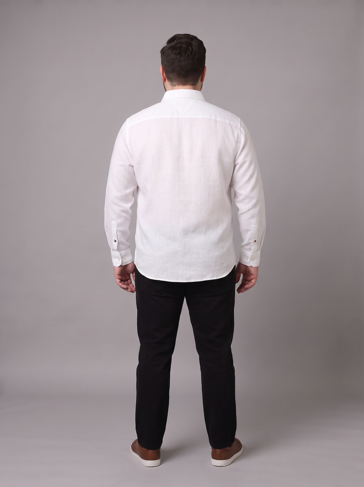 A man viewed from behind wearing a white long-sleeve button-up shirt, black trousers, and brown shoes, standing against a plain gray studio background.