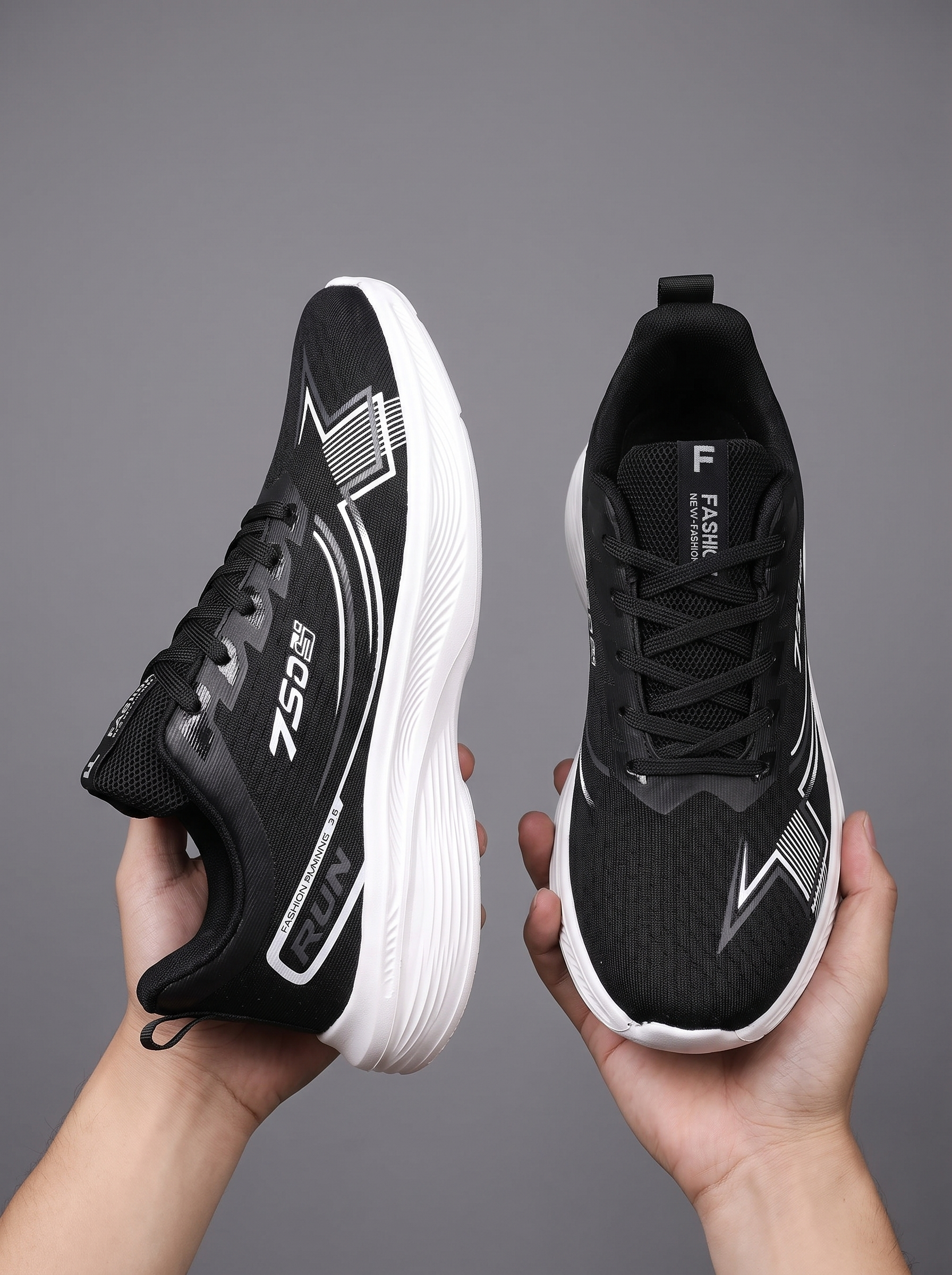 A pair of black and white athletic sneakers held up by two hands against a solid gray studio background. The shoes feature a breathable mesh upper, black laces, and a thick white cushioned sole.