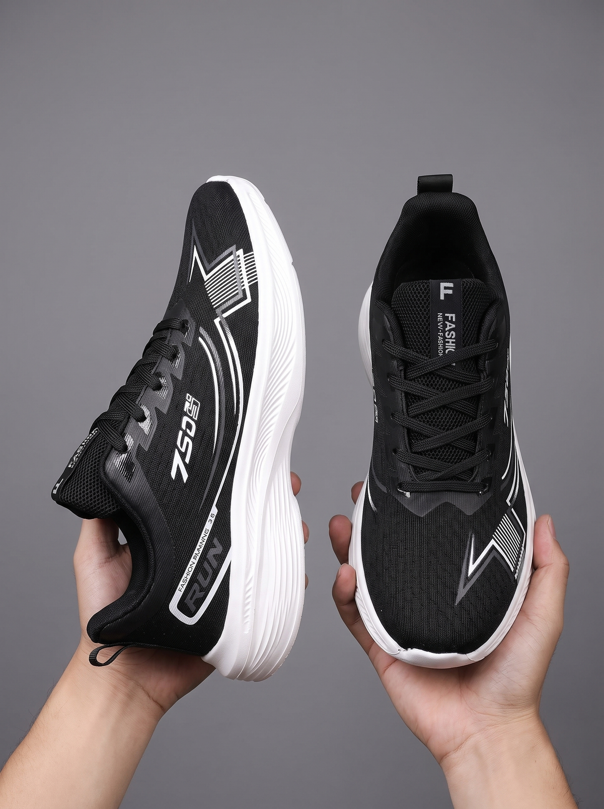 A pair of black and white athletic sneakers held up by two hands against a solid gray studio background. The shoes feature a breathable mesh upper, black laces, and a thick white cushioned sole.