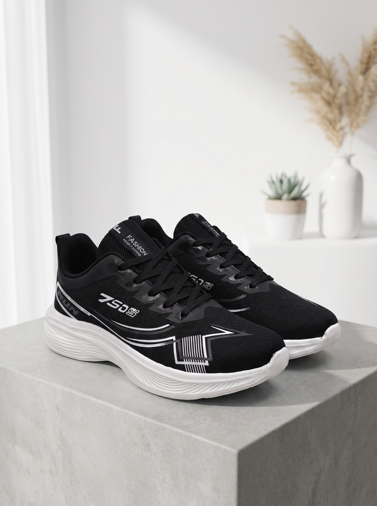 A pair of black and white athletic sneakers displayed on a light gray concrete block with a blurred indoor background featuring a small potted succulent and a vase.