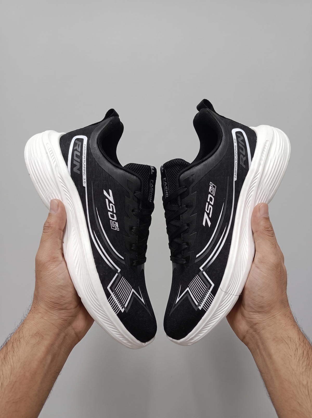 A pair of black and white athletic sneakers held up by two hands against a plain gray background. The shoes feature a black mesh upper with white graphic accents and thick white soles.