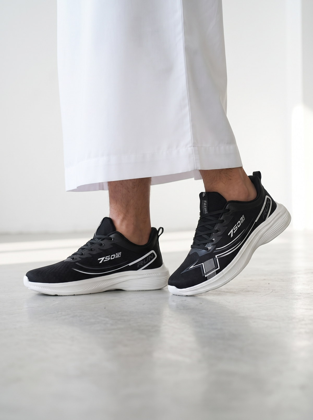 Black and white athletic sneakers worn by a person in white trousers, shown from the knees down in a bright indoor setting.