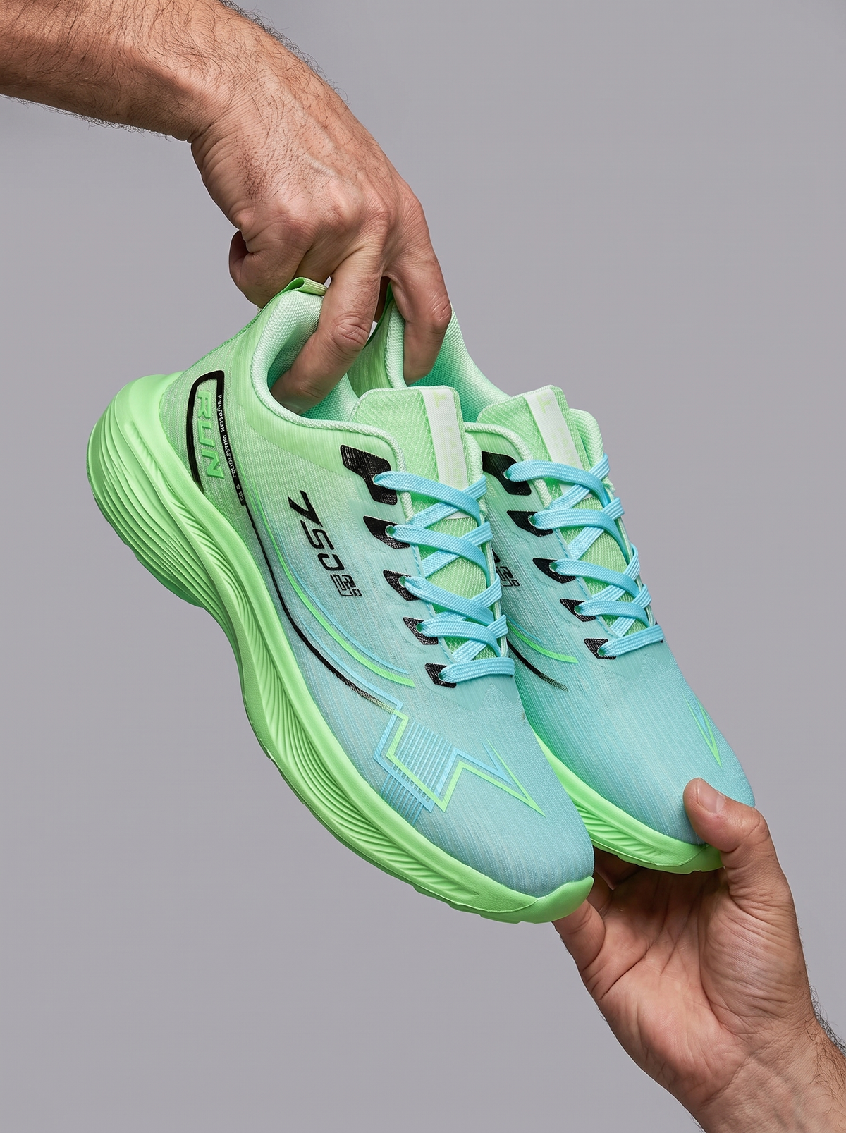 A pair of light green and mint blue running shoes held up by two hands against a solid gray background. The shoes feature a textured mesh upper, light blue laces, and black branding details.
