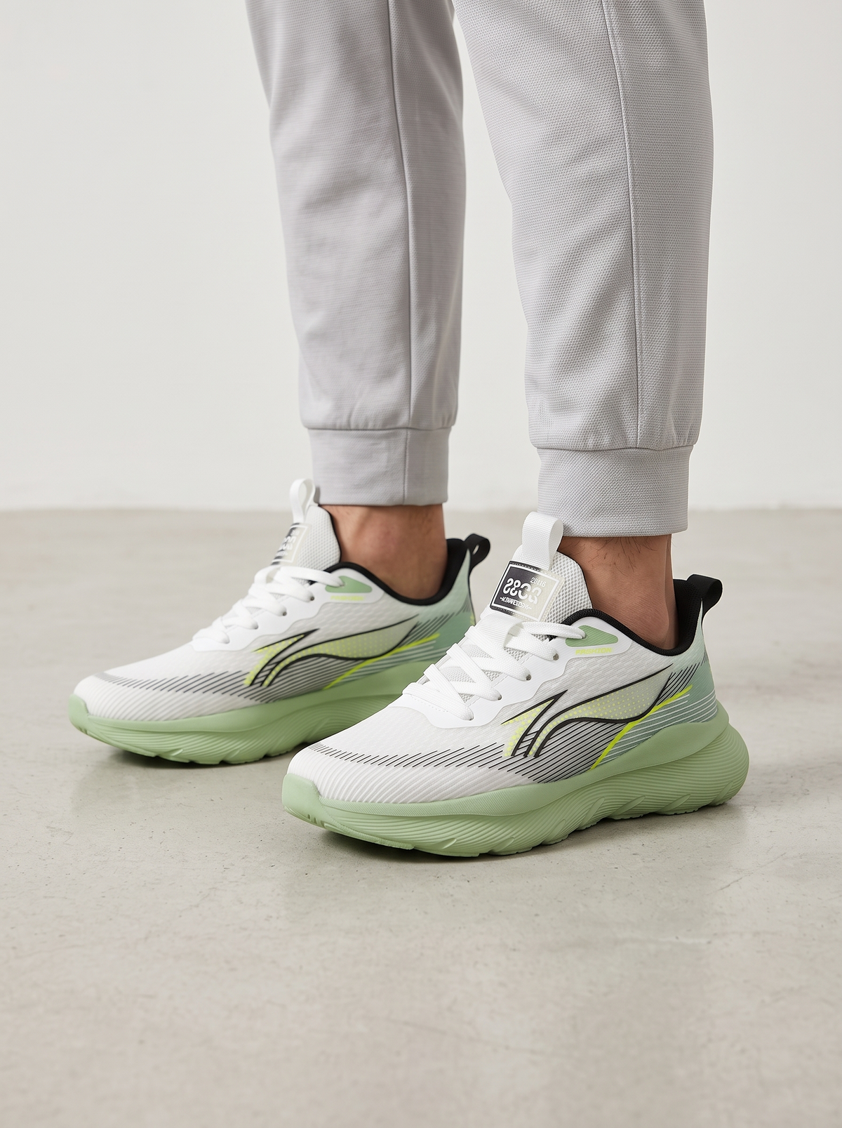 White athletic sneakers with mint green soles and gray and lime green accents, shown on a model wearing light gray joggers against a neutral studio background.