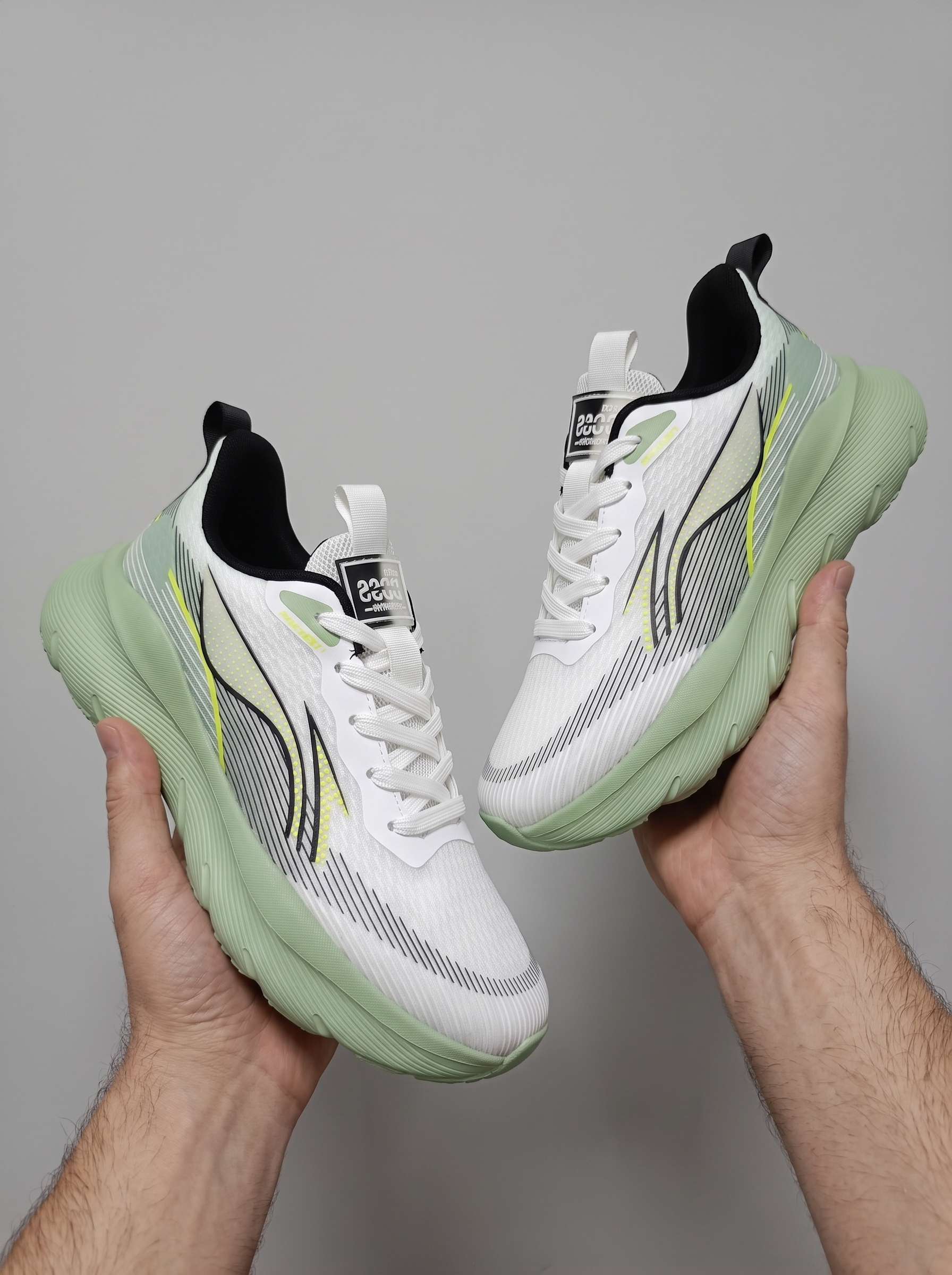 A pair of white athletic sneakers with mint green soles and light gray side stripes, held up by two hands against a plain gray background.