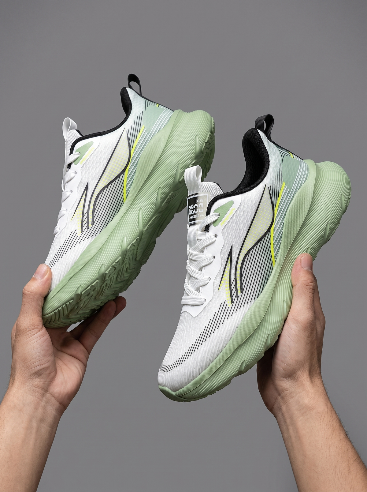 A pair of white and mint green athletic sneakers held up by two hands against a solid gray background.