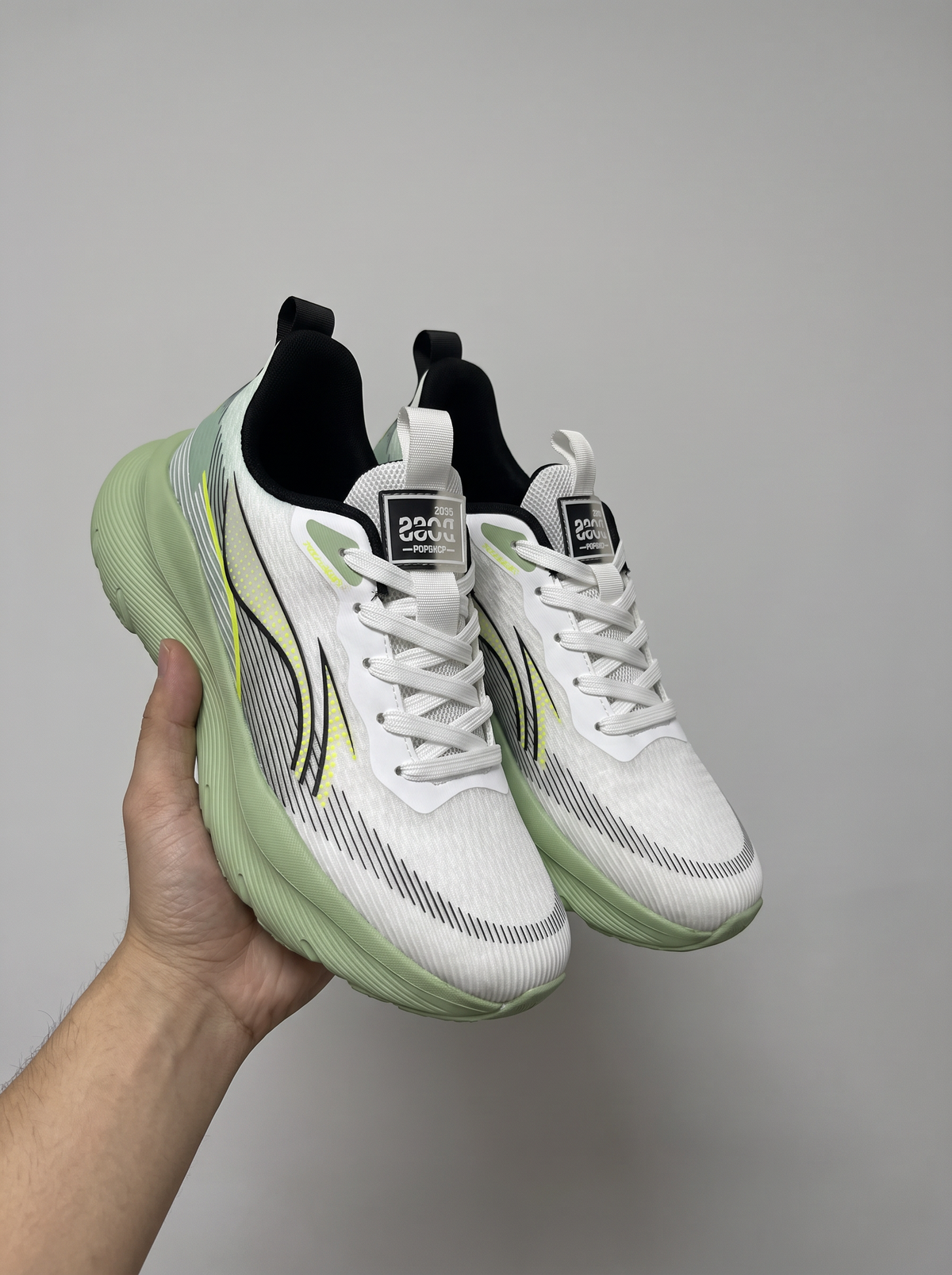 A pair of white running shoes with mint green soles and black accents, held up by a hand against a plain gray background.
