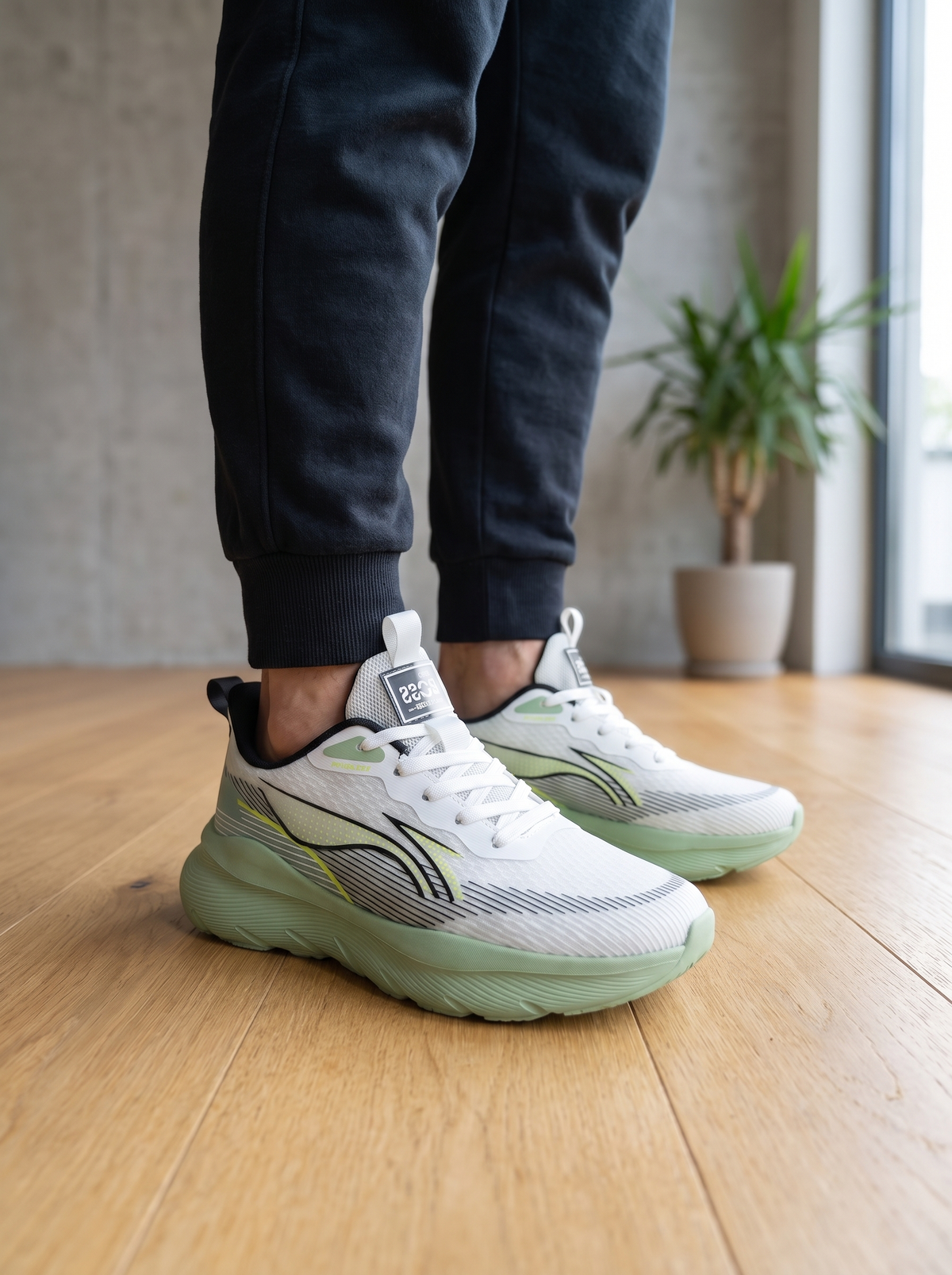 White athletic sneakers with light green soles and black-and-green side stripes, shown on a person wearing dark joggers in an indoor room with a wood floor and potted plant.