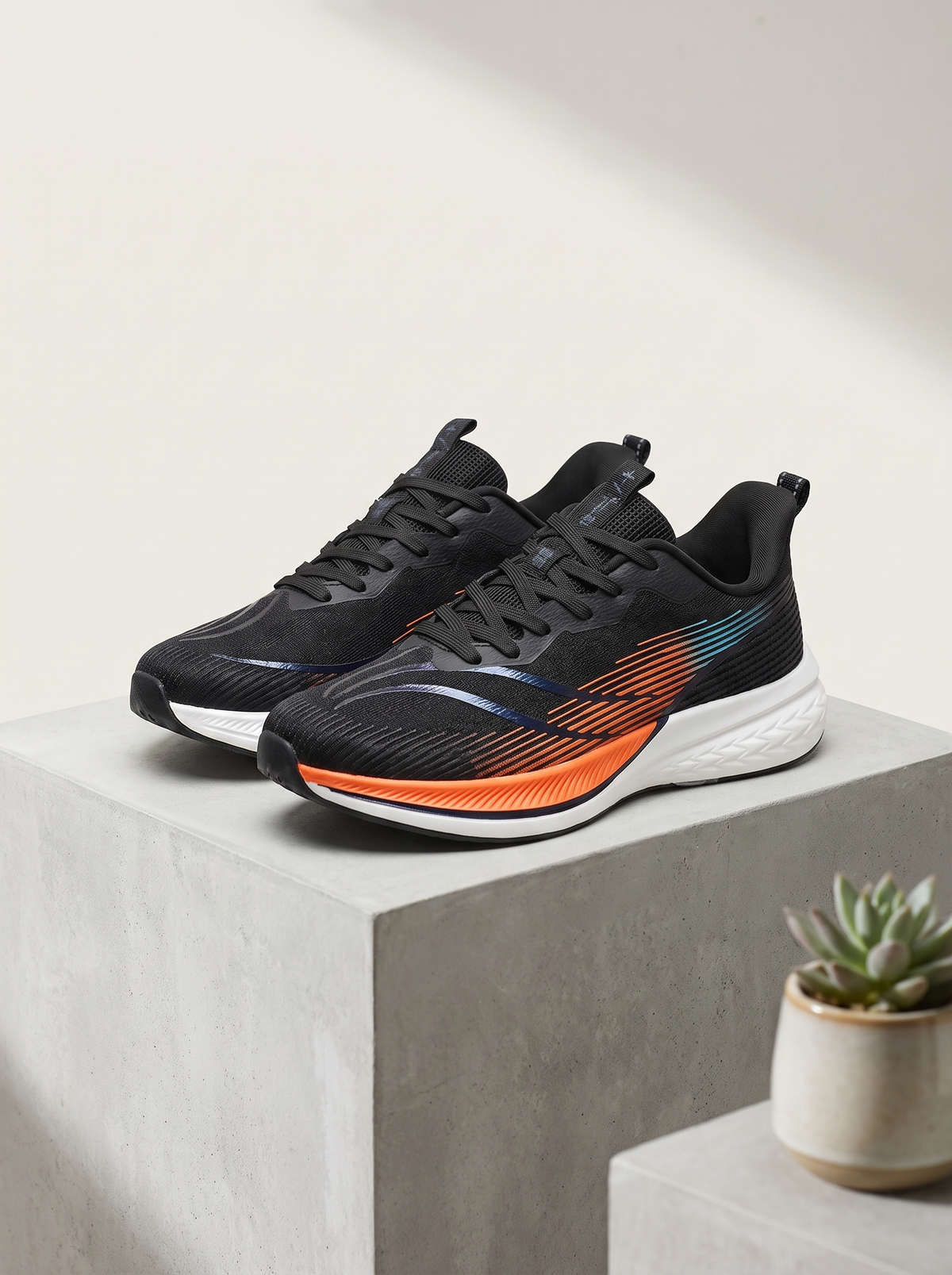 A pair of black athletic running shoes with vibrant orange and teal accents on the midsole, displayed on a light gray concrete pedestal with a small potted succulent in the background.