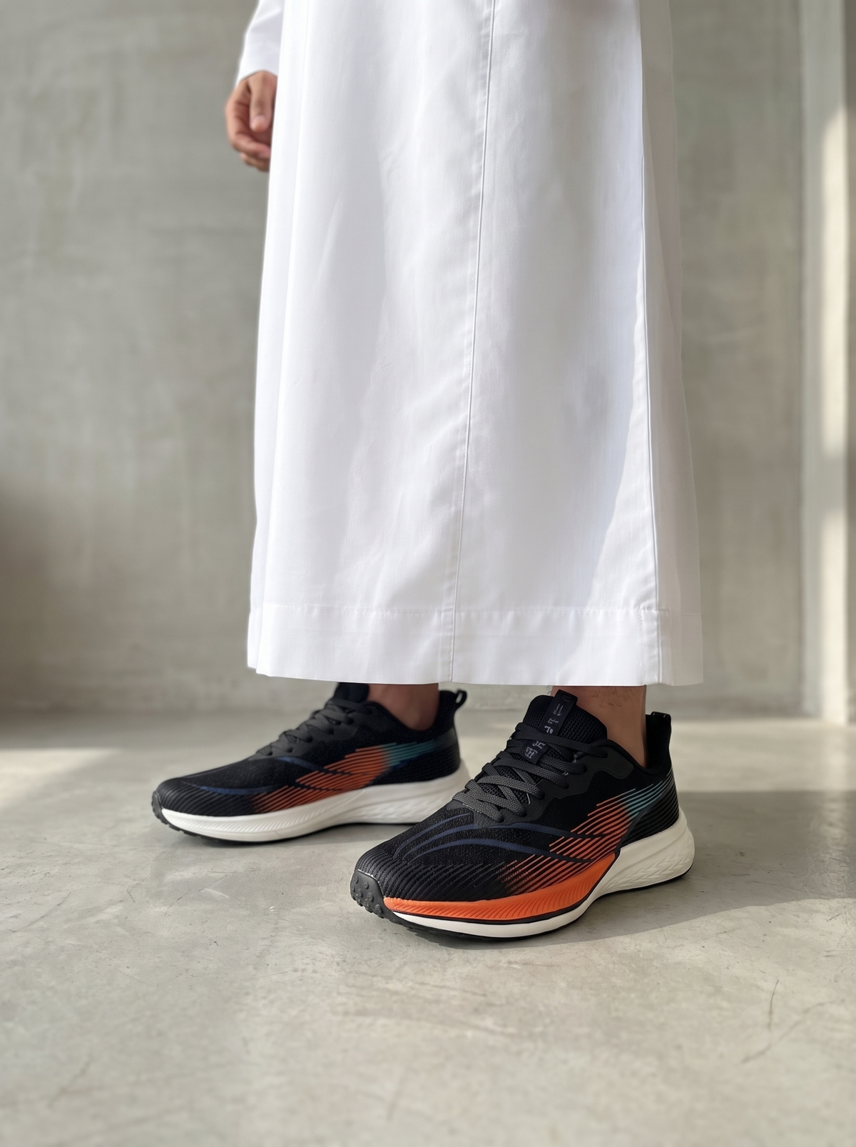 A person wearing a long white robe stands indoors on a light concrete floor, showing a pair of black athletic sneakers with orange and teal accents.