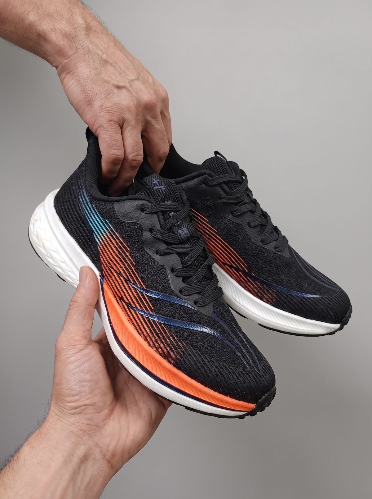 A pair of black running shoes with vibrant orange and blue accents, held by two hands against a neutral gray background.