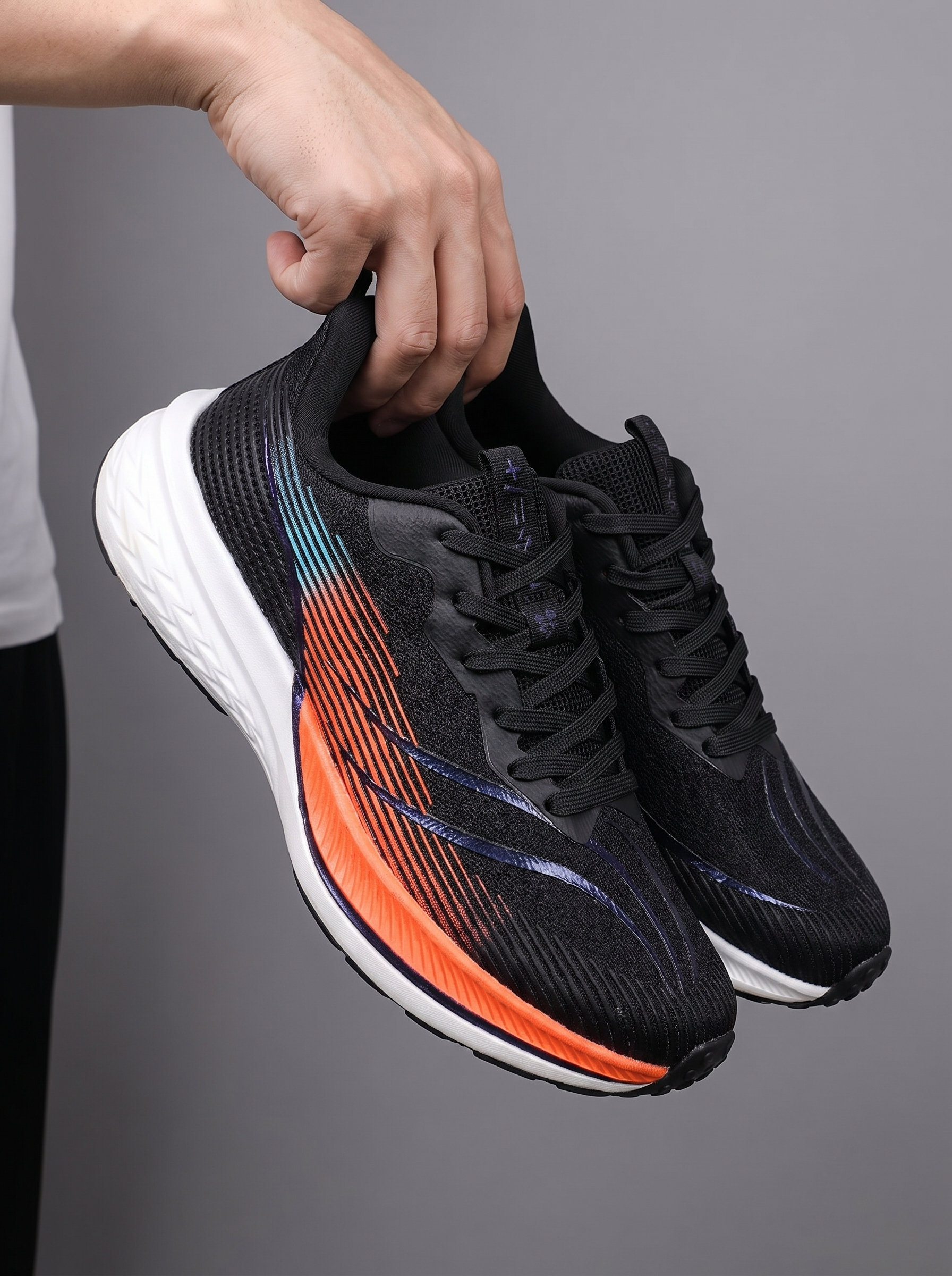 A person's hand holding a pair of black athletic running shoes with vibrant orange, blue, and purple gradient accents on the side, set against a solid gray background.