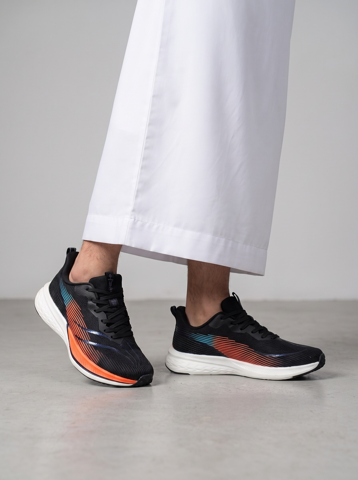 A person wearing black athletic sneakers with orange and teal accents, paired with white wide-leg trousers, standing on a light gray floor against a gray wall.