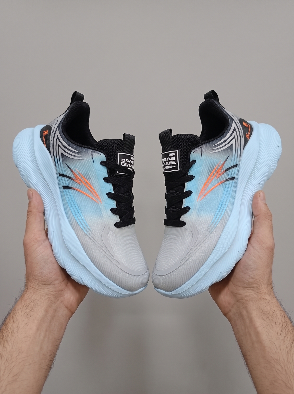 A pair of Reebok sneakers held up by two hands against a plain gray background. The sneakers feature a light gray and white mesh upper with a blue-to-light-blue gradient, black laces, and orange Reebok logos.