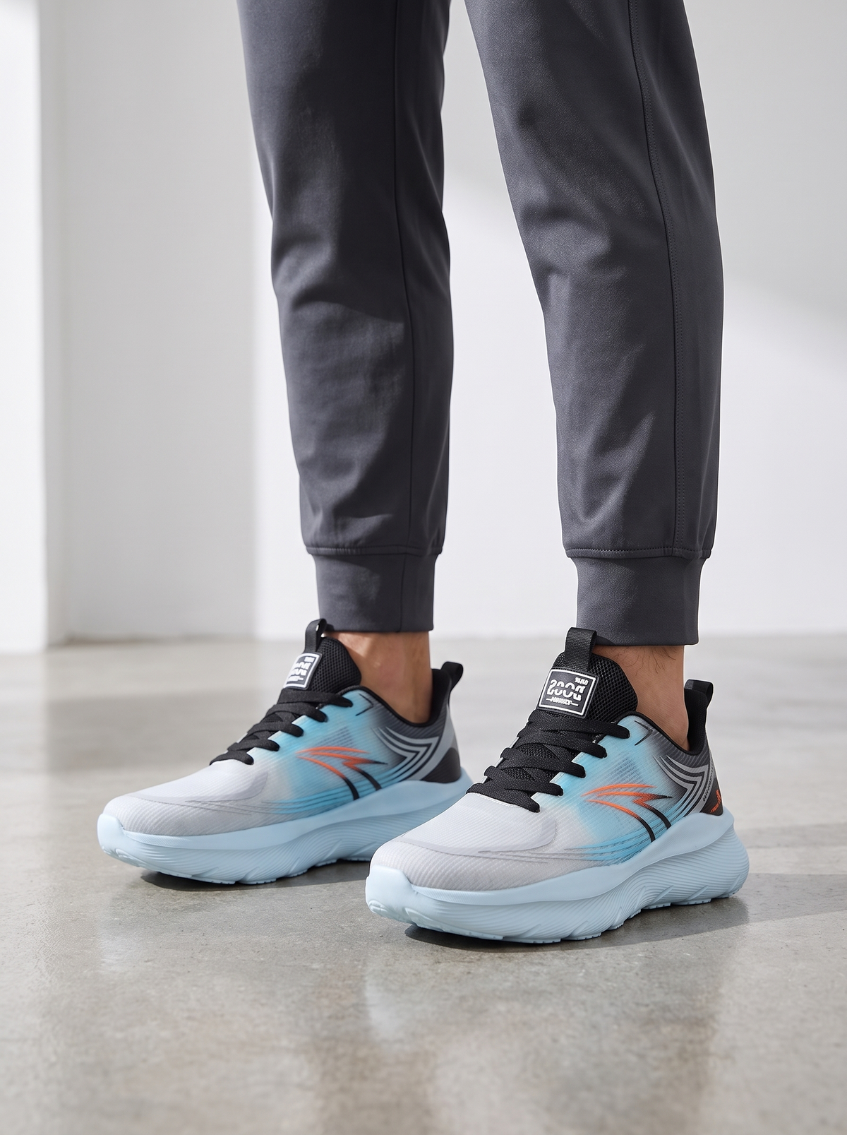 A person wearing gray jogger pants and Reebok sneakers with a light blue, gray, and black design and orange accents, standing on a concrete floor.