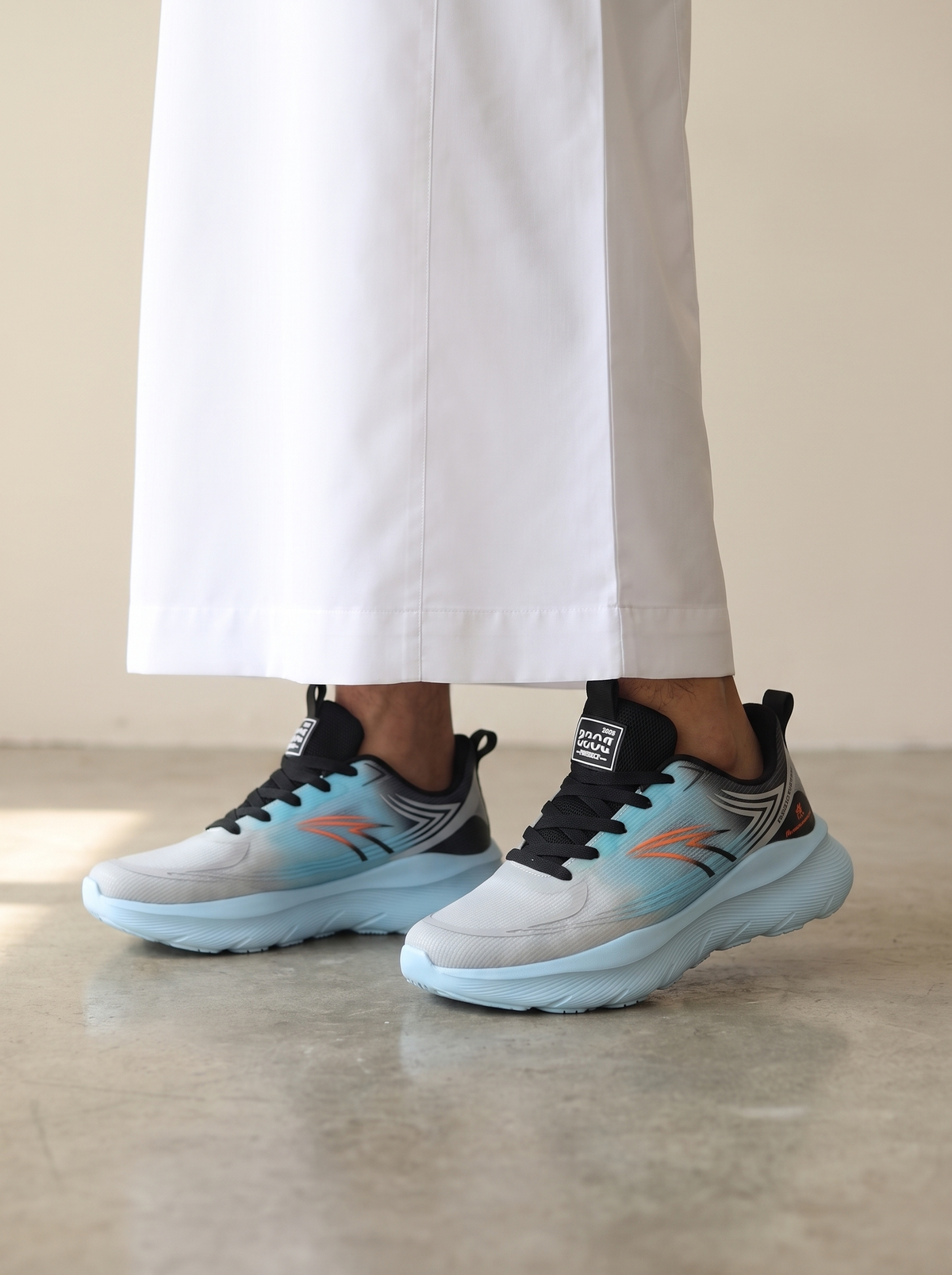 A person wearing light gray and light blue running shoes with black laces and orange accents, paired with white wide-leg pants, standing on a concrete floor.