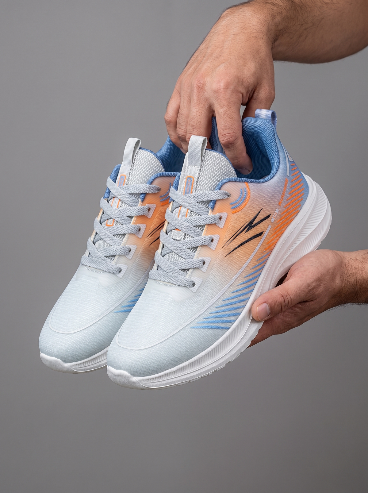 A pair of athletic running shoes held up by two hands against a solid gray studio background. The shoes feature a white and light blue gradient upper with orange gradient accents and a blue collar lining.