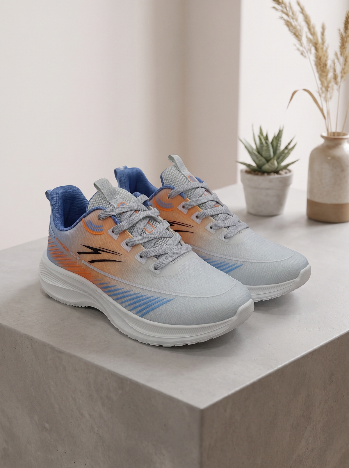 A pair of light gray running shoes with orange and blue accents, displayed on a light gray stone pedestal with a small potted succulent and dried grass in a vase in the background.