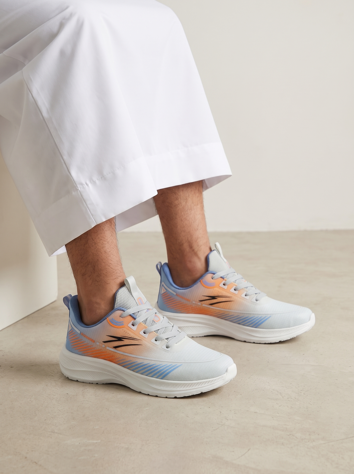 A person wearing white draped pants and light blue and orange Reebok sneakers, seated on a beige floor against a neutral beige wall.
