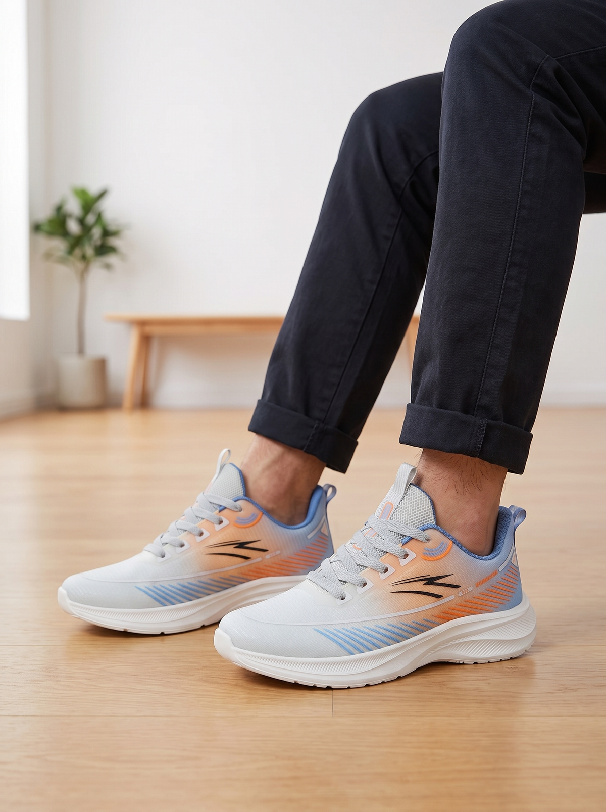 A person wearing light gray athletic sneakers with orange and blue accents, paired with dark navy pants, sitting on a wooden floor in a bright indoor room.