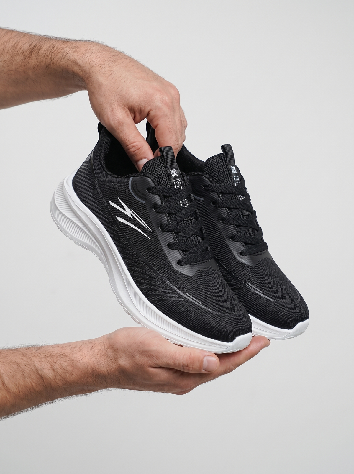 A pair of black athletic sneakers with white soles and white logo accents, held by a person's hands against a light gray background.