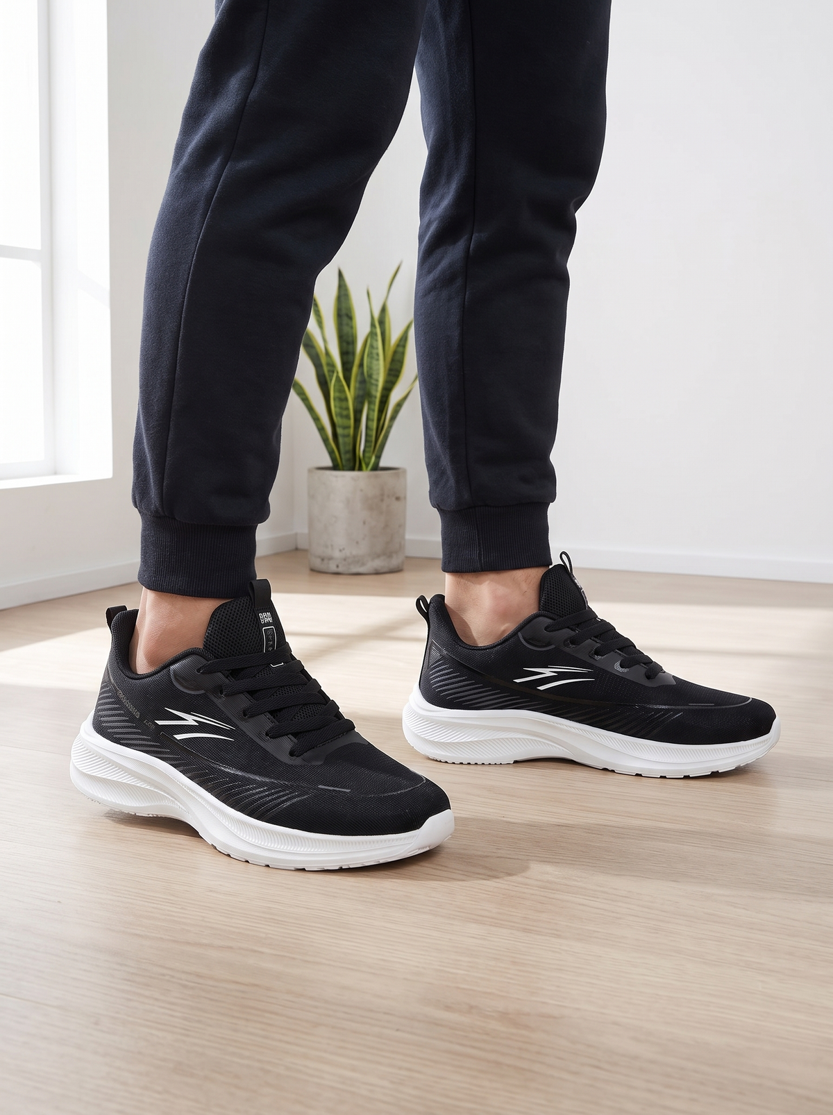 A person wearing black athletic sneakers with white soles and dark jogger pants, standing on a light wood floor in a bright indoor room with a small potted plant in the background.