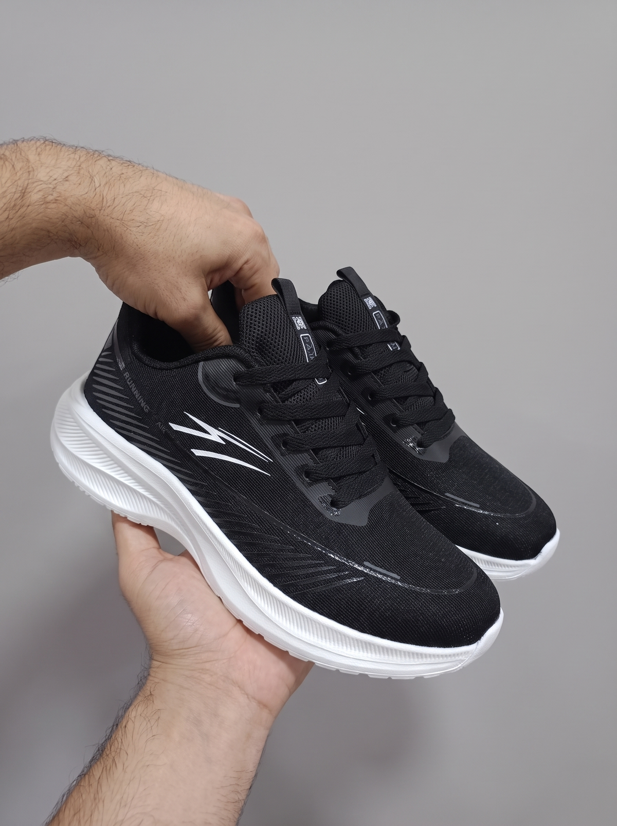 A pair of black athletic sneakers with white soles, held in a person's hands against a plain gray background.