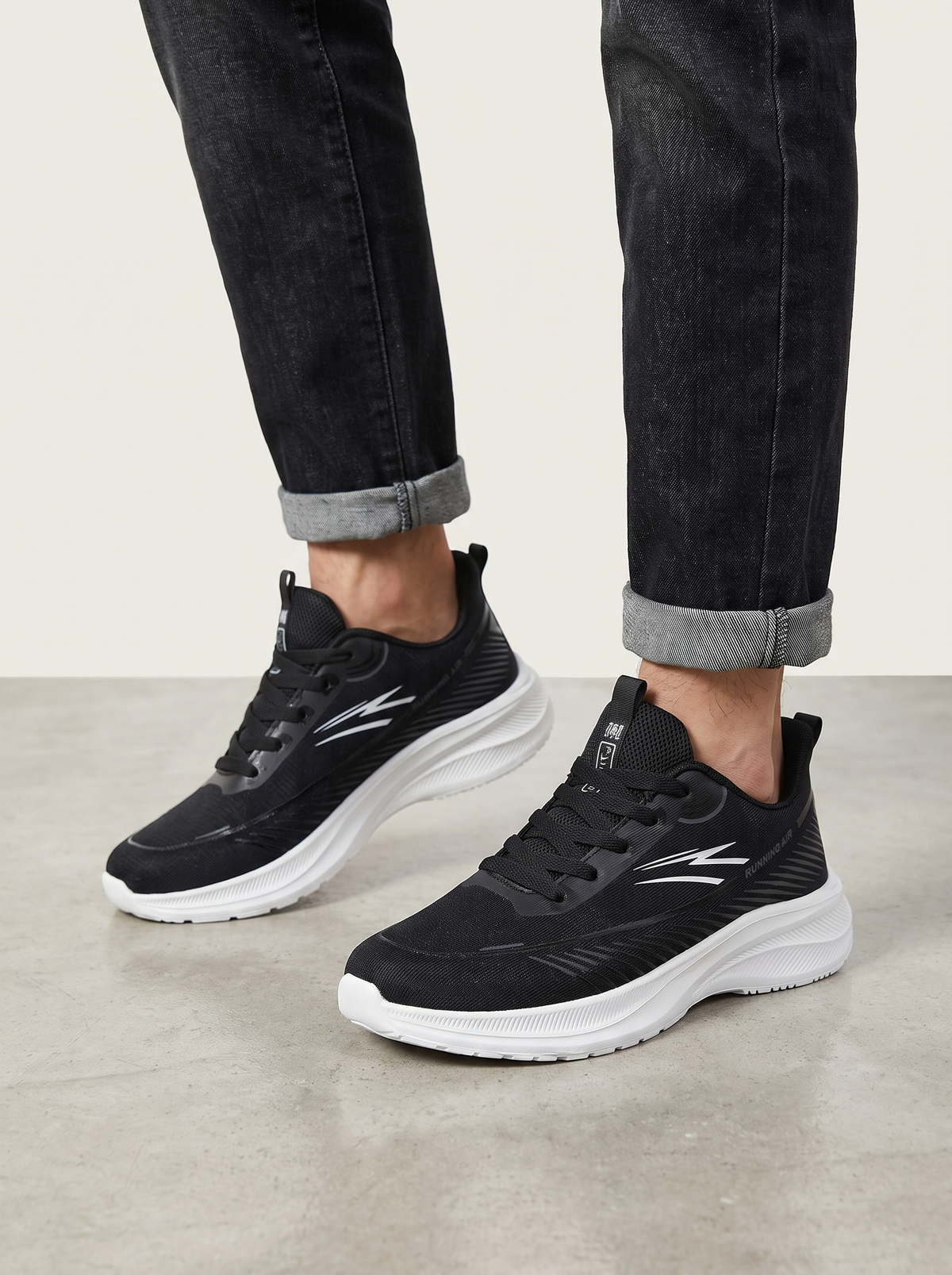 Black athletic sneakers with white chunky soles and silver logo details, shown on a model wearing dark cuffed jeans against a light gray studio background.