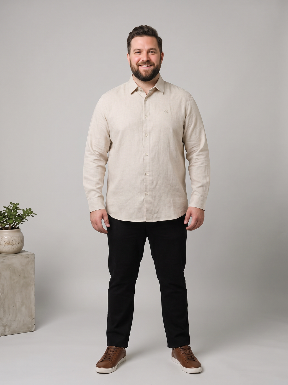 A man wearing a light beige linen button-down shirt, black trousers, and brown shoes, standing against a neutral gray studio background with a small potted plant on a concrete block to the left.