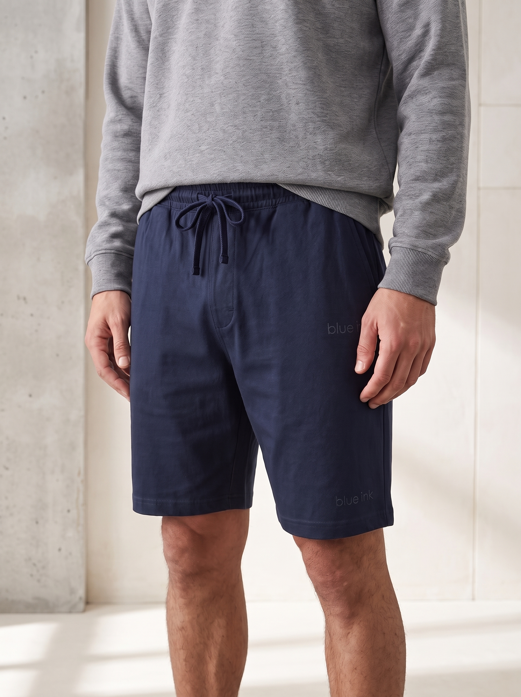 A man wearing navy blue drawstring shorts and a gray sweatshirt, standing in a brightly lit indoor setting with a concrete wall.