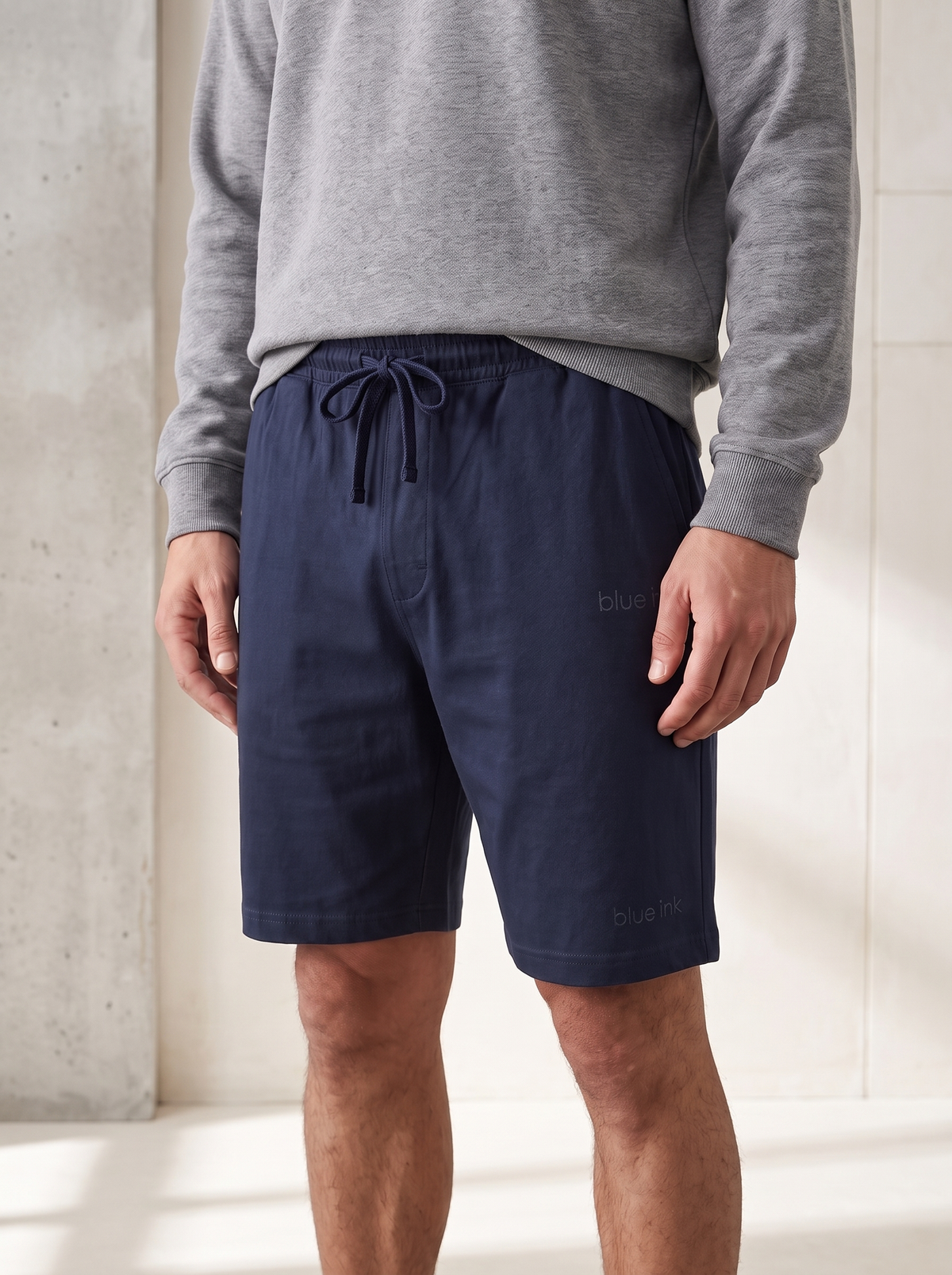 A man wearing navy blue drawstring shorts and a gray sweatshirt, standing in a brightly lit indoor setting with a concrete wall.