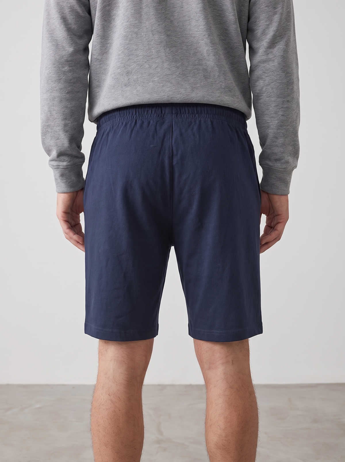 A man viewed from the back wearing navy blue shorts and a gray long-sleeve sweatshirt, standing against a plain light gray wall.